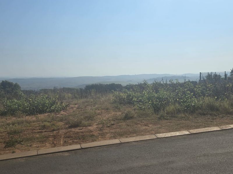 A view of a vacant residential plot featuring natural vegetation, paved road access, and distant landscape vistas.