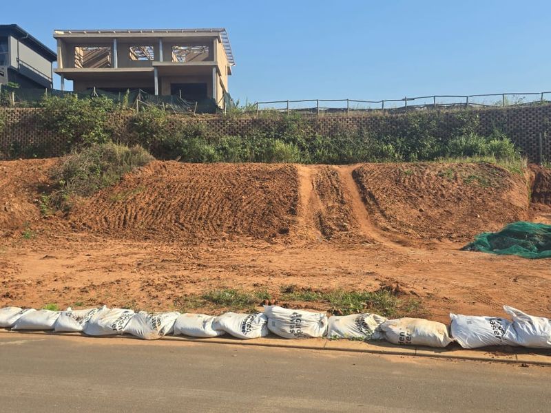 A vacant residential plot featuring a sloped terrain with a retaining wall, currently undergoing site preparation with sandbags along the street boundary.