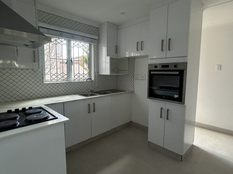 A clean, modern kitchen featuring white cabinetry, built-in oven, electric hob, extractor fan, and a tiled backsplash with a window overlooking the exterior.