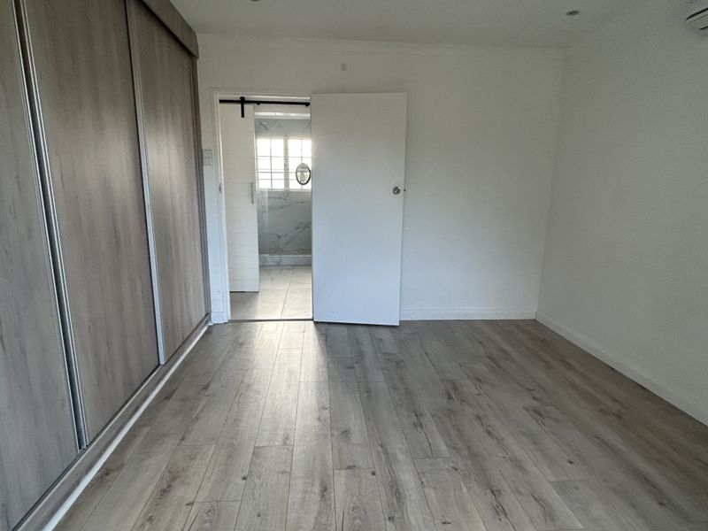 A bright, empty bedroom featuring modern laminate flooring, a large built-in sliding wardrobe, and direct access to an en-suite bathroom.