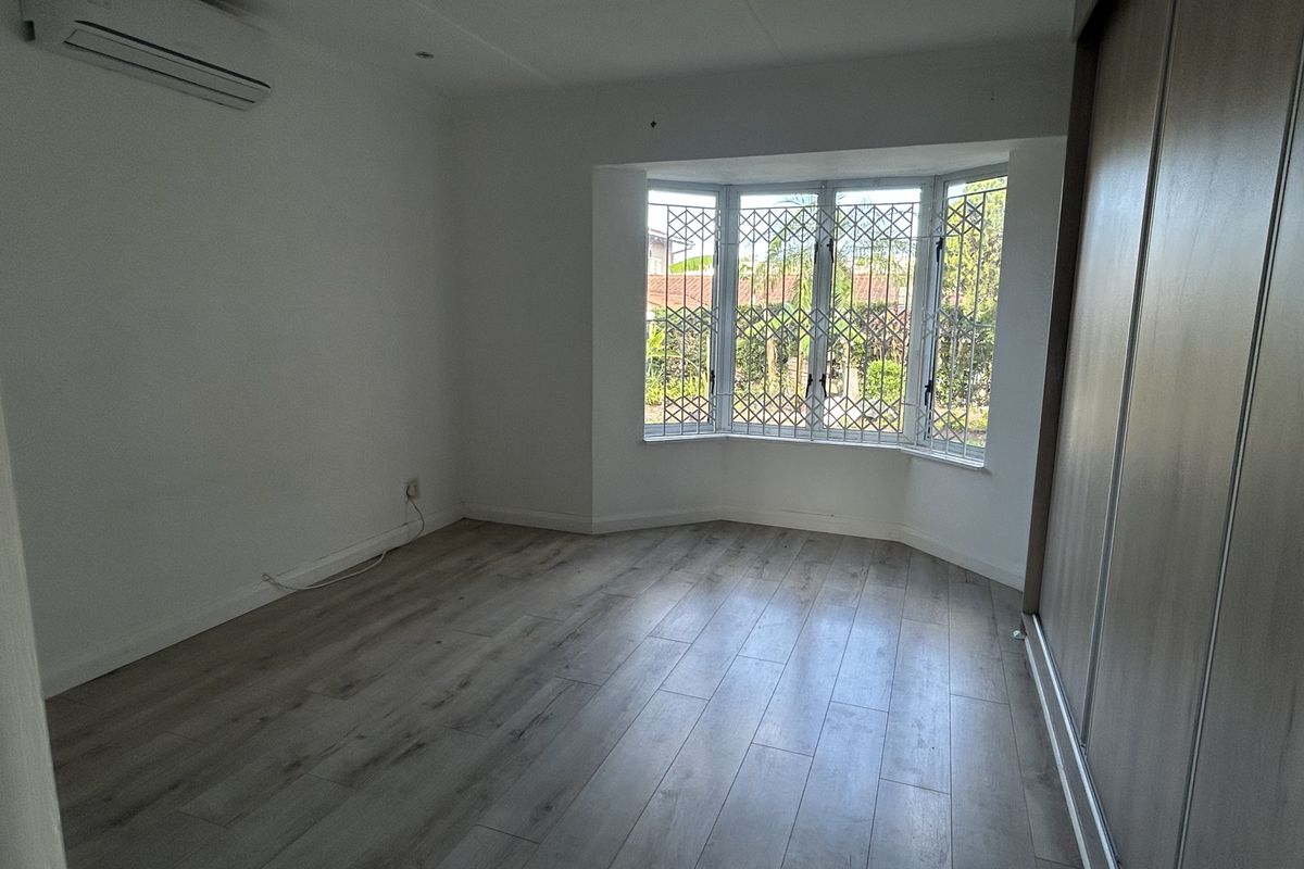 A bright, empty bedroom featuring laminate flooring, a built-in wardrobe, an air conditioning unit, and a bay window with security bars.