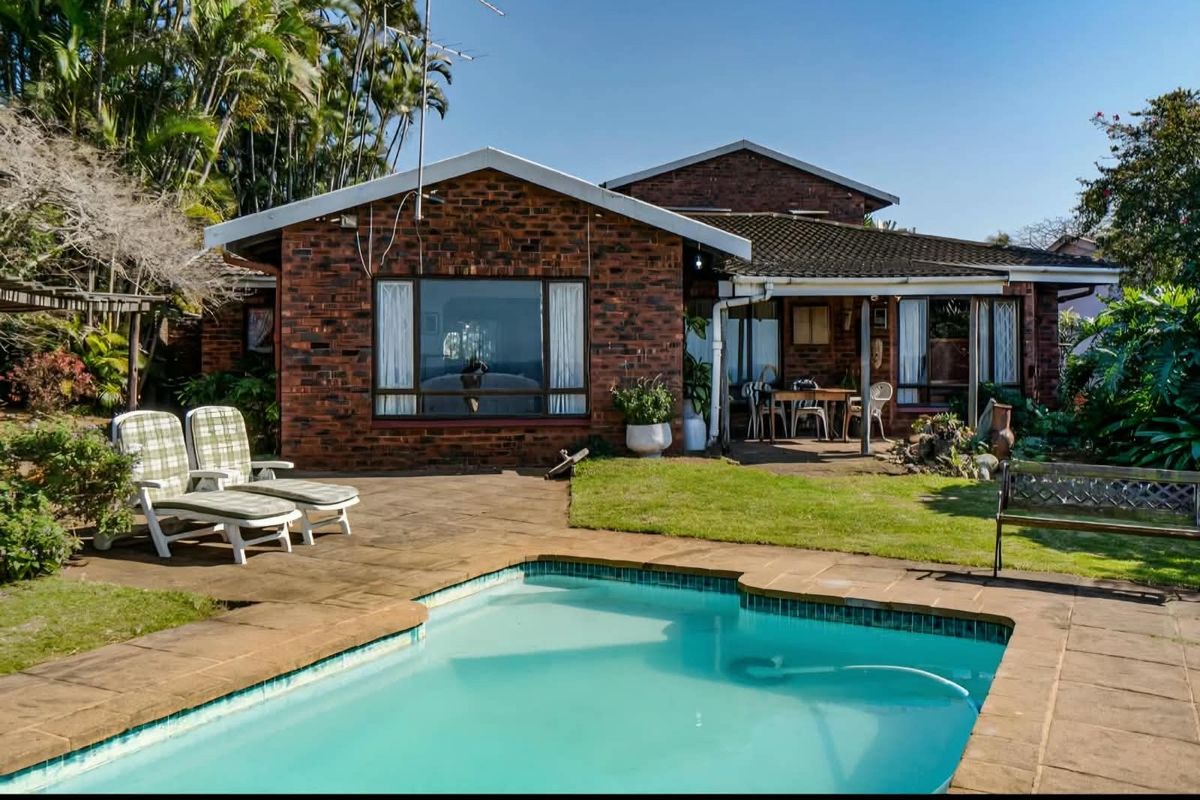 Rear view of a brick house featuring a swimming pool, paved deck area, and a covered patio overlooking the garden.