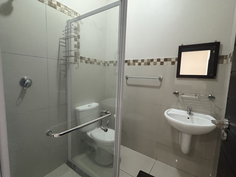 A functional bathroom featuring a glass-enclosed shower, pedestal washbasin, and toilet with neutral wall tiling and chrome fixtures.