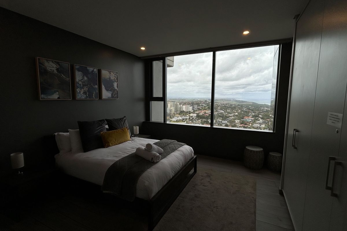 Modern bedroom featuring dark accent walls, built-in cabinetry, and expansive floor-to-ceiling windows offering panoramic city and coastal views.