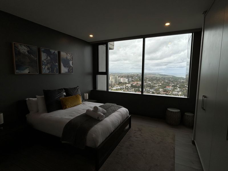 Modern bedroom featuring dark accent walls, built-in cabinetry, and expansive floor-to-ceiling windows offering panoramic city and coastal views.