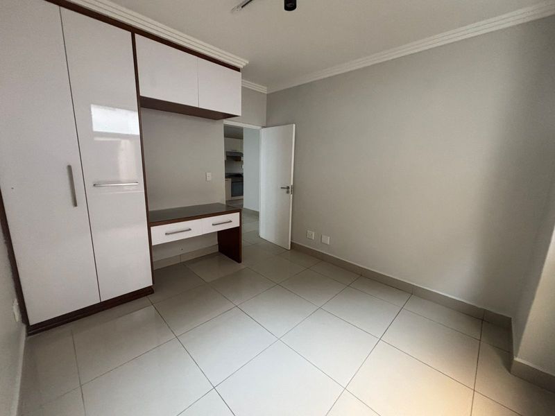 A bright, empty bedroom featuring modern built-in cupboards with an integrated desk, neutral tiled flooring, and clean finishes throughout.