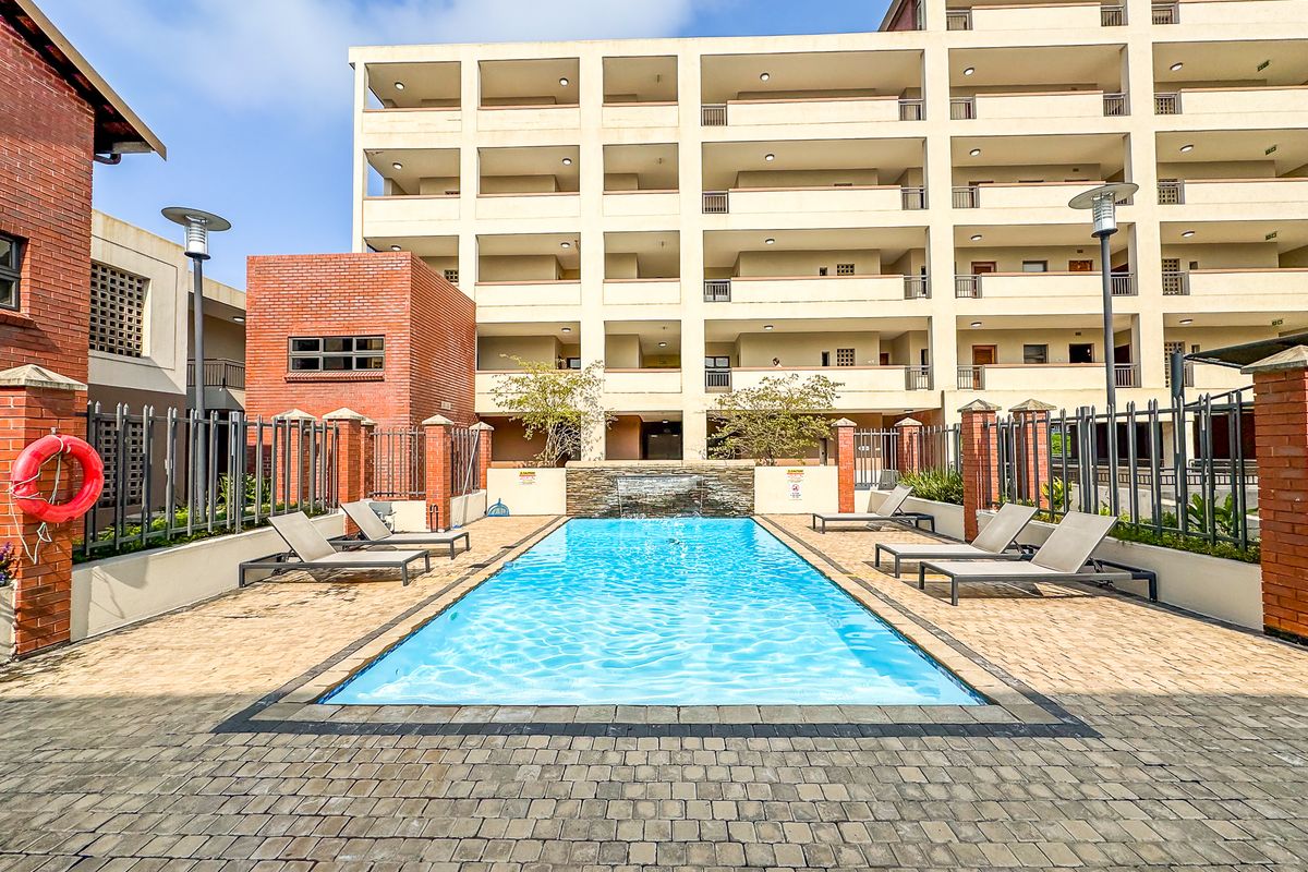 A well-maintained communal swimming pool area featuring a water feature, paved decking, and secure metal fencing within an apartment complex.