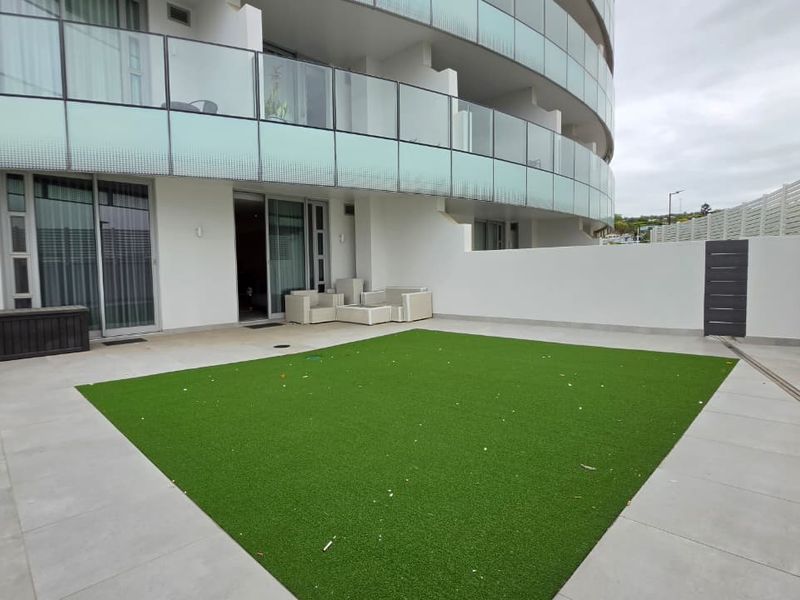 A spacious ground-floor patio featuring tiled flooring, artificial turf, and a modern glass balustrade, providing direct access to the apartment interior.