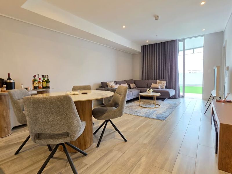 Bright open-plan living and dining area featuring wood-look tile flooring, recessed ceiling lights, and a sliding glass door leading to an outdoor space.
