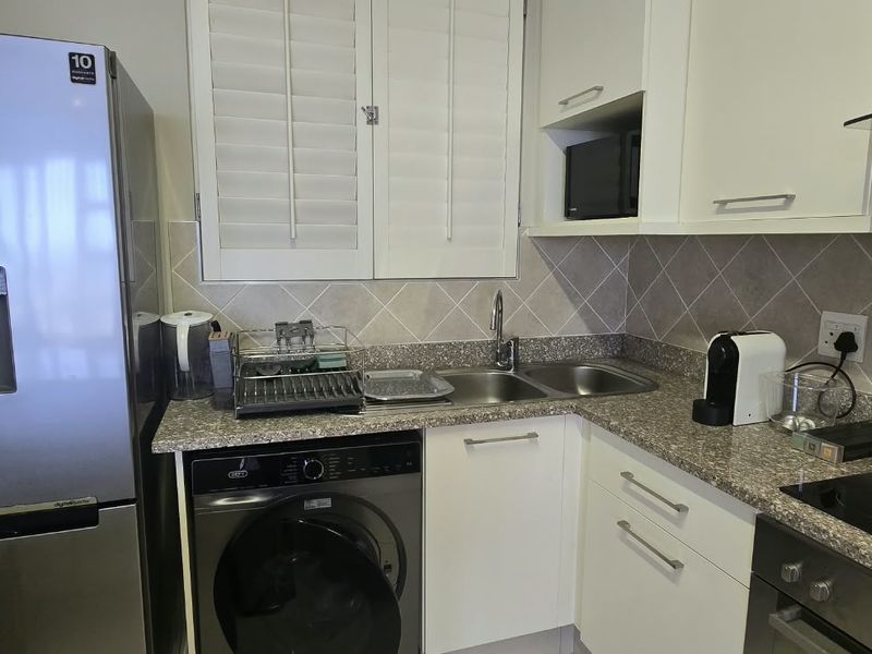 A functional kitchen featuring granite countertops, a double bowl sink, built-in oven with electric hob, extractor fan, and white cabinetry with shutter blinds.
