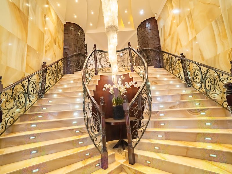 A grand entrance hall featuring a sweeping dual staircase with ornate wrought iron balustrades, marble steps with integrated lighting, and elegant stone wall finishes.