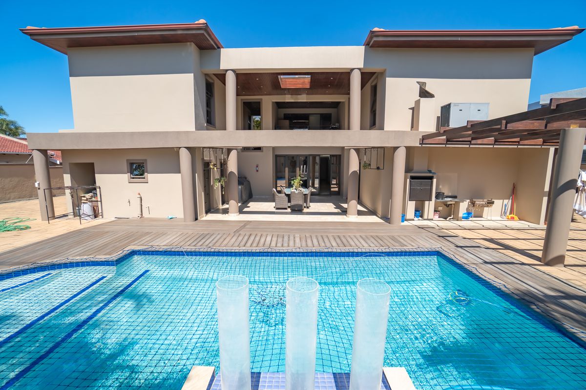 Rear elevation featuring a swimming pool with safety net, timber decking, and a spacious covered patio area with built-in braai and pergola.