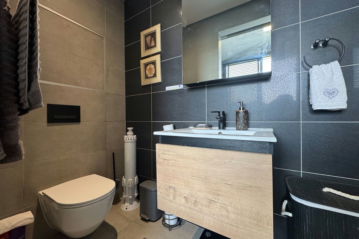 Modern bathroom featuring a wall-mounted toilet with concealed cistern, a wood-finish vanity cabinet with integrated basin, and dark tiled walls.