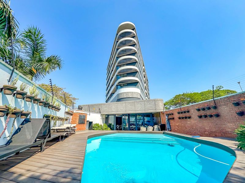 A well-maintained outdoor pool area featuring timber decking, brick boundary walls with electric fencing, and a view of the modern apartment building facade.