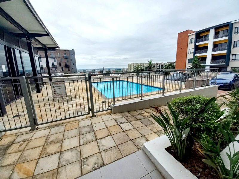 Communal swimming pool area featuring paved patio space, metal safety fencing, and landscaped garden planters within an apartment complex setting.