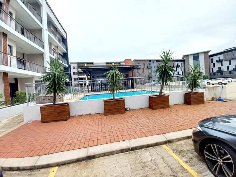 A well-maintained communal swimming pool area featuring paved surrounds, metal safety fencing, and modern apartment building architecture in the background.