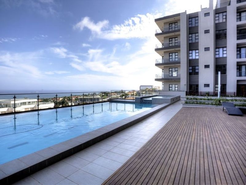 A modern rooftop swimming pool area featuring timber decking, glass balustrades, and panoramic coastal views set against a contemporary apartment building.