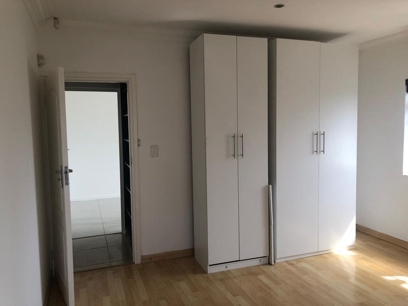 A bright, empty bedroom featuring light laminate flooring, white built-in cupboards, and recessed ceiling lighting, with an open doorway leading to an adjacent room.