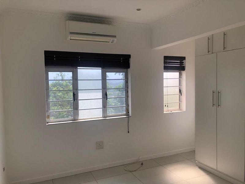 A bright, empty bedroom featuring tiled floors, built-in cupboards, an air conditioning unit, and security bars on the windows.