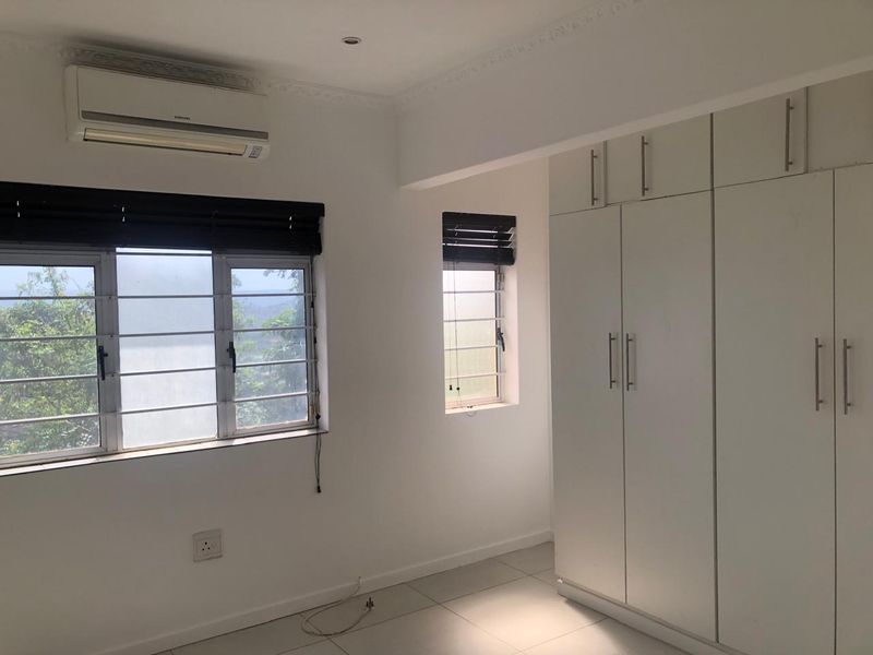 A bright, empty bedroom featuring tiled floors, built-in cupboards, and a wall-mounted air conditioning unit with security bars on the windows.