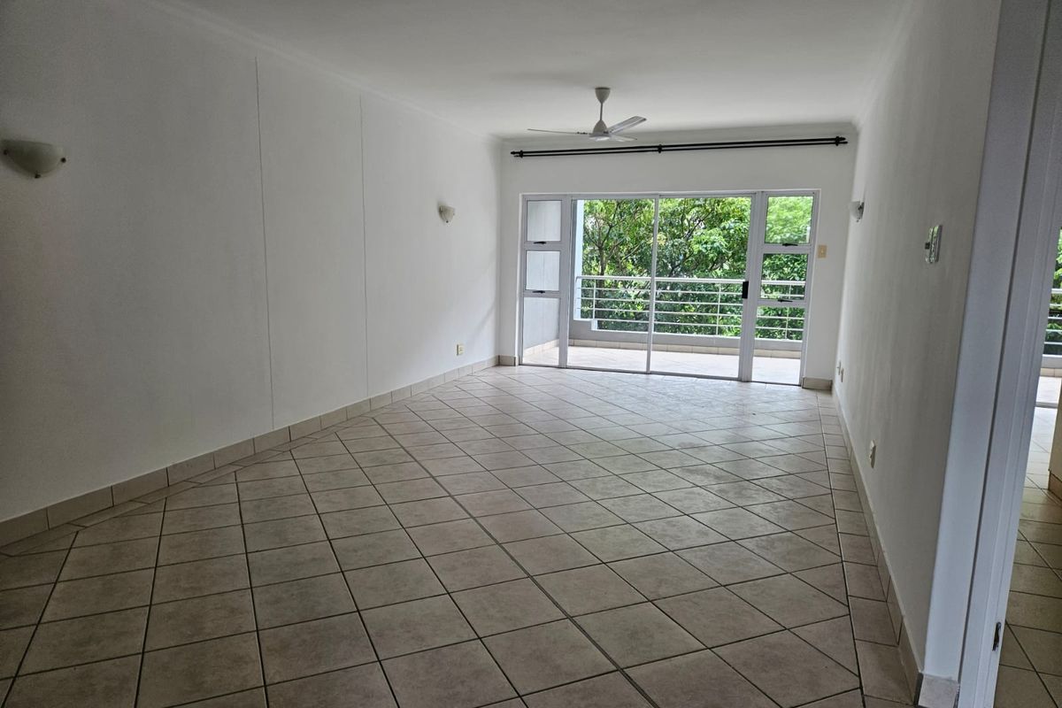 Spacious, empty living area featuring neutral tiled flooring, a ceiling fan, and large sliding glass doors opening onto a balcony with leafy views.