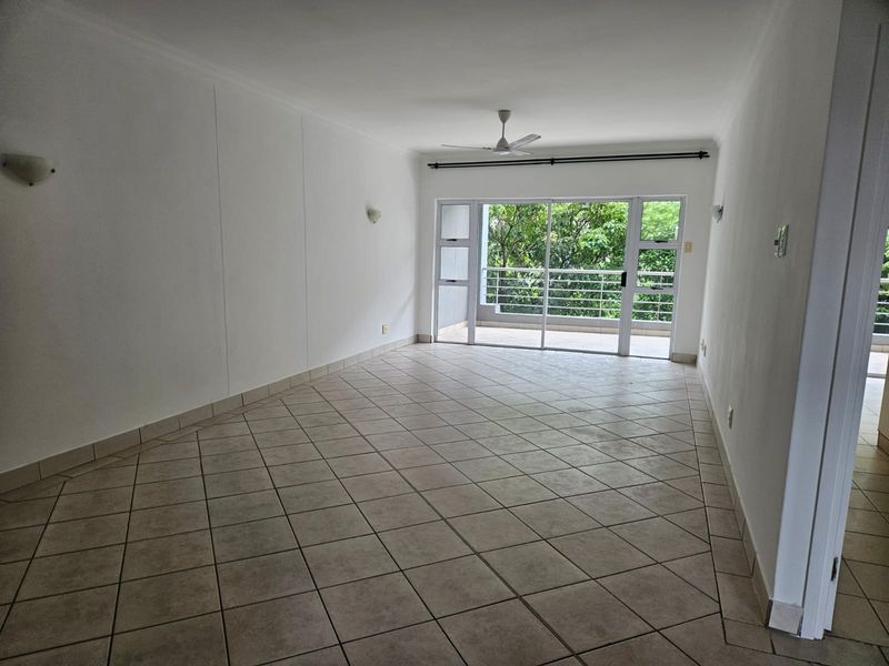 Spacious, empty living area featuring neutral tiled flooring, a ceiling fan, and large sliding glass doors opening onto a balcony with leafy views.