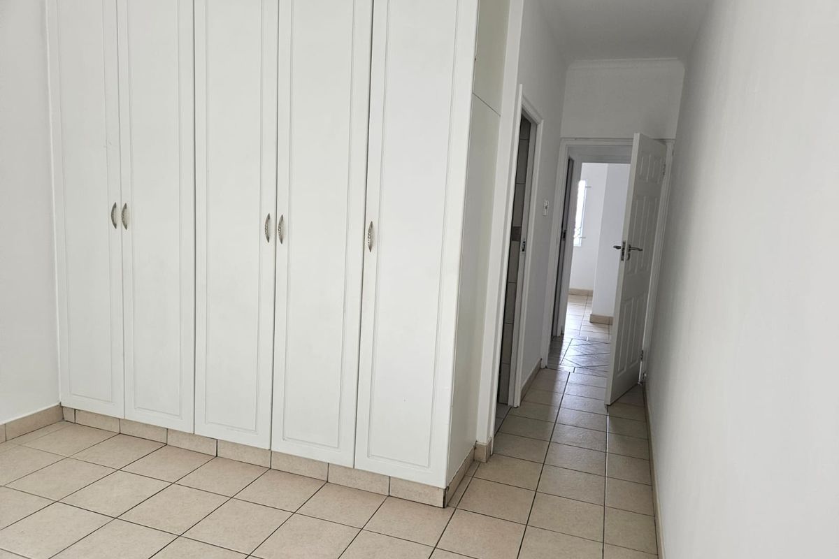 A clean, empty bedroom featuring white built-in cupboards and neutral tiled flooring, leading into a hallway with access to other rooms.