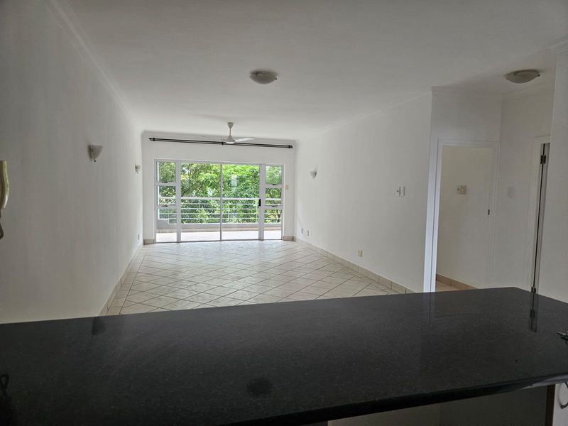 Spacious open-plan living area featuring tiled flooring, a granite breakfast bar, and large sliding glass doors leading to a balcony with greenery views.