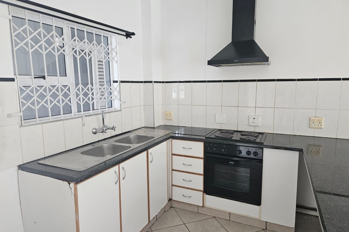 A functional kitchen featuring granite countertops, built-in oven, electric hob, extractor fan, and double sink with tiled backsplash and security window bars.