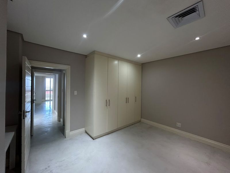 A bright, empty bedroom featuring polished concrete floors, built-in cupboards, and recessed lighting, with a view into the adjacent hallway.