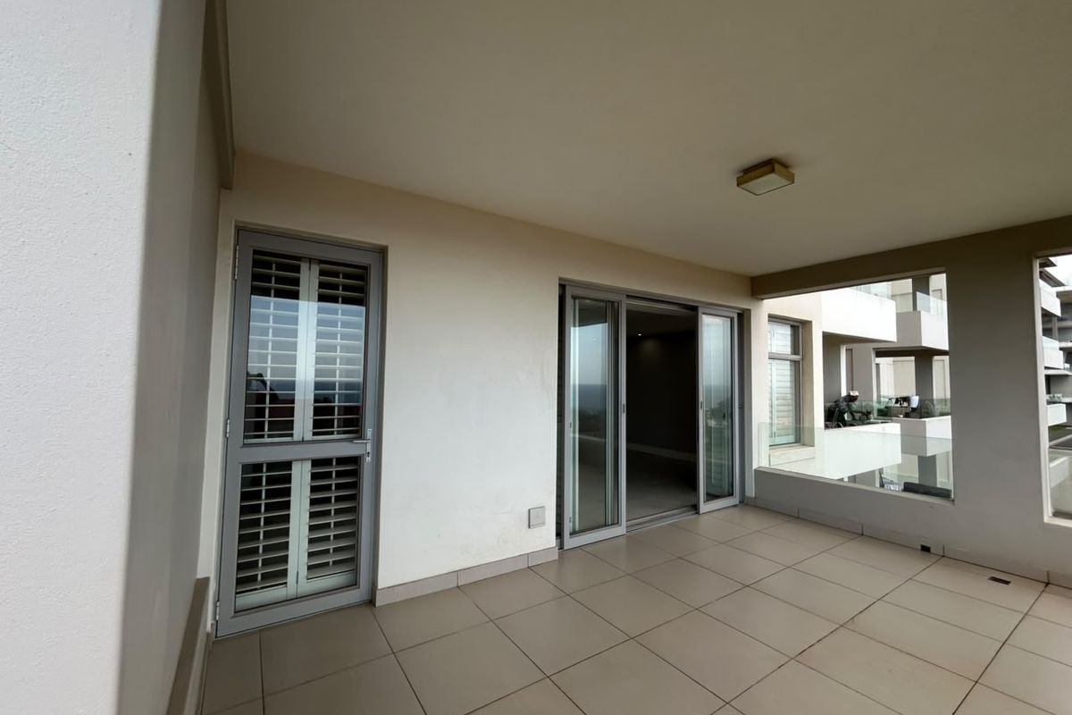 A spacious covered balcony featuring tiled flooring, a glass balustrade, and sliding door access to the interior living area.