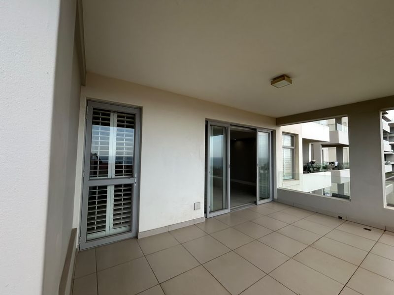 A spacious covered balcony featuring tiled flooring, a glass balustrade, and sliding door access to the interior living area.