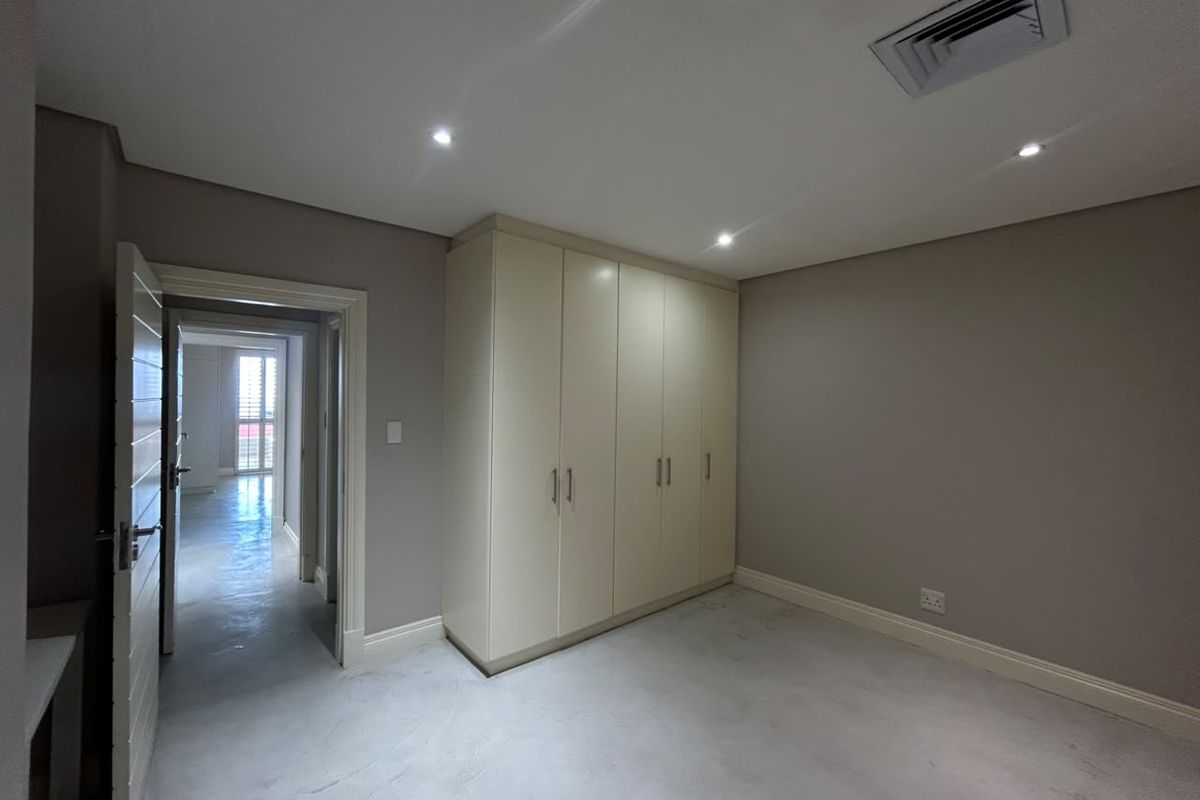 A bright, empty bedroom featuring polished concrete floors, built-in cupboards, and recessed lighting, with a view into the adjacent hallway.