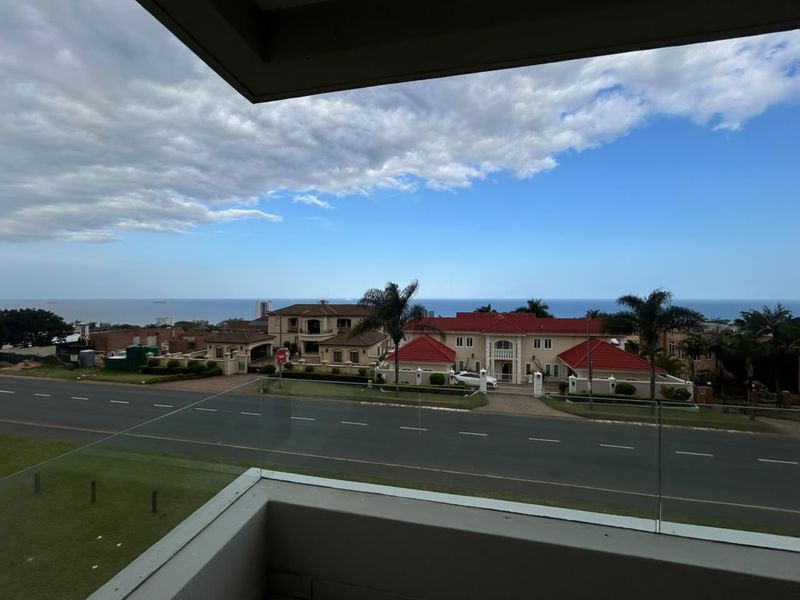 A scenic elevated view overlooking a residential street and the ocean, framed by a modern glass balcony balustrade.