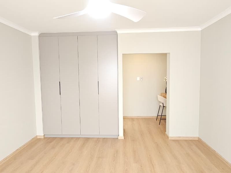 A bright, neutral bedroom featuring modern built-in cupboards, light laminate flooring, and a ceiling fan, offering a clean and versatile living space.