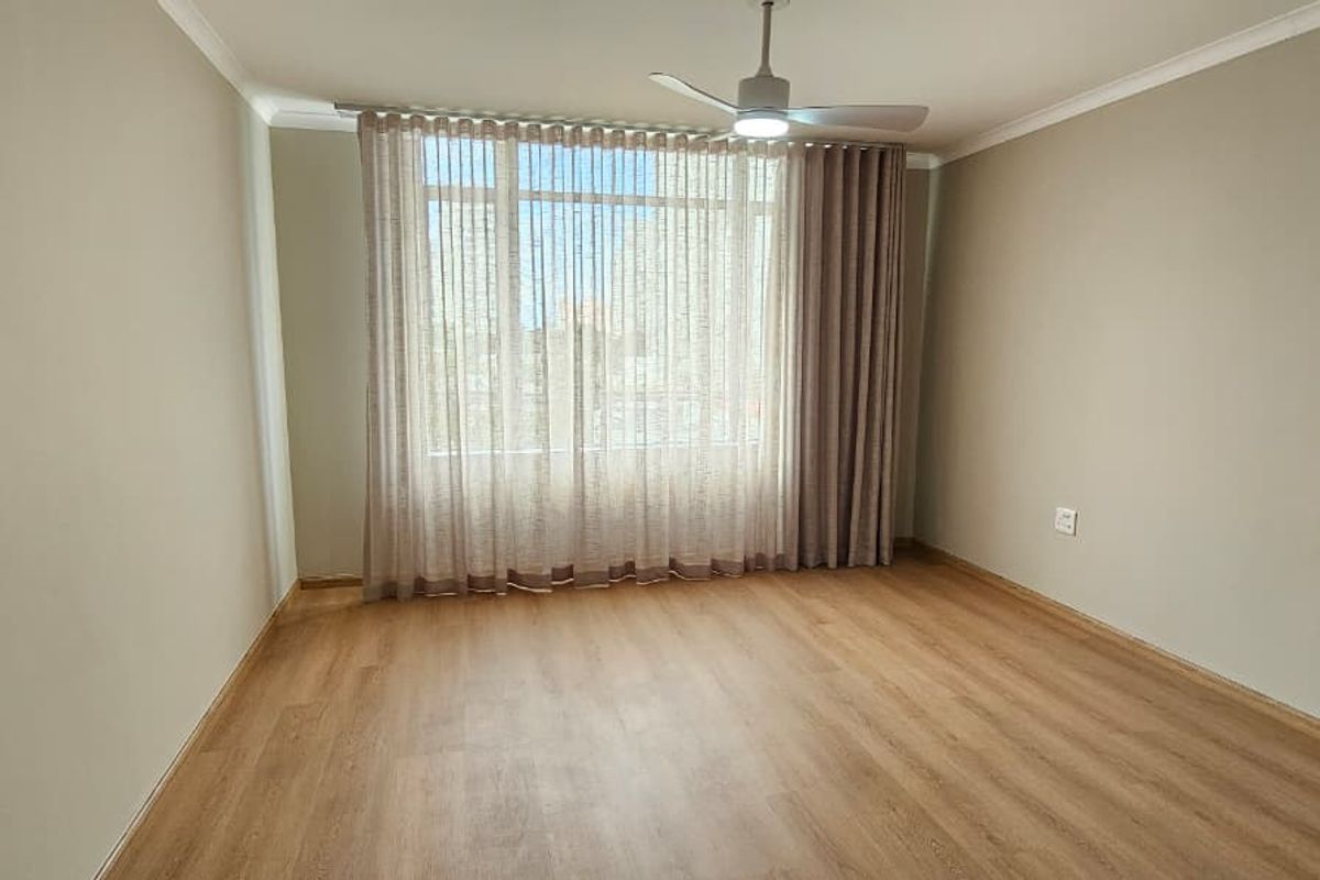 A bright and spacious empty bedroom featuring light-toned laminate flooring, a ceiling fan, and large windows with neutral curtains.