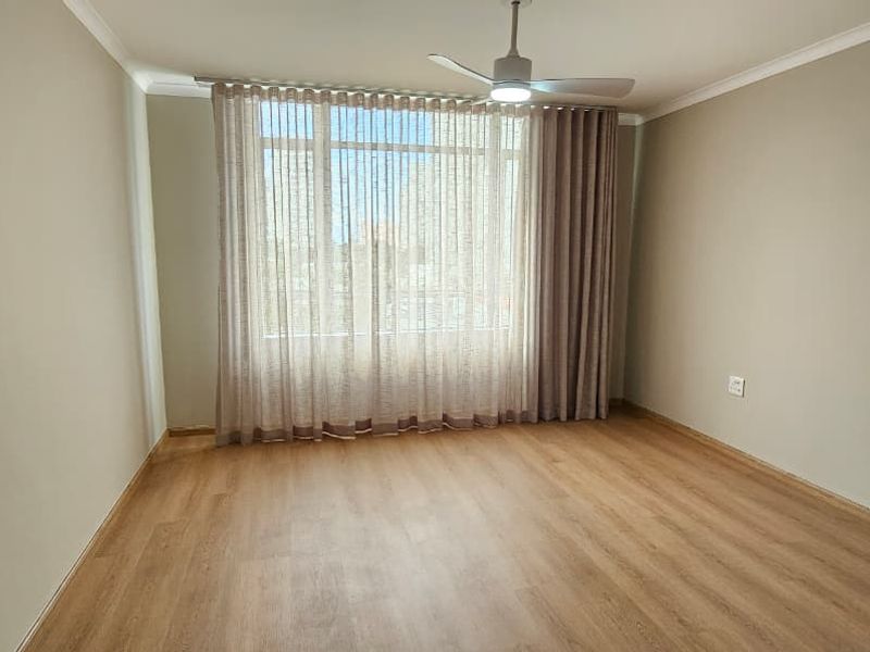 A bright and spacious empty bedroom featuring light-toned laminate flooring, a ceiling fan, and large windows with neutral curtains.