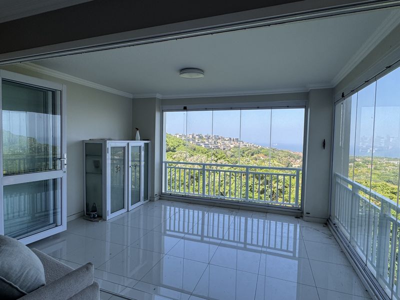 A bright, enclosed balcony featuring tiled flooring, a white metal balustrade, and expansive glass panels offering scenic hillside and coastal views.