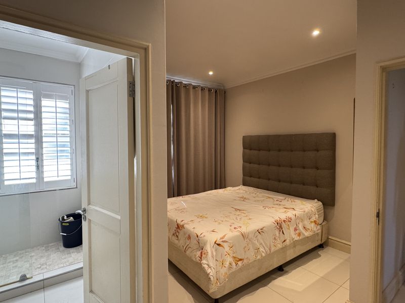 A well-maintained bedroom featuring tiled flooring, recessed downlighting, and plantation shutters, with an adjacent bathroom visible through the doorway.