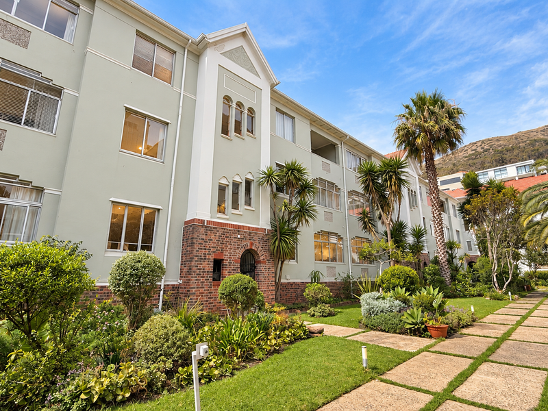 A well-maintained apartment building exterior featuring a brick and plaster facade, large windows, and a landscaped garden with a paved walkway.