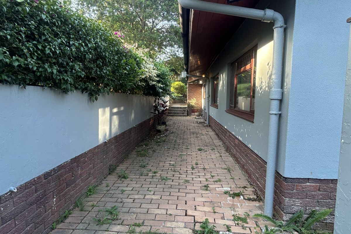 A narrow side passage featuring brick paving, a boundary wall with hedge, and external drainage infrastructure along the side of the house.