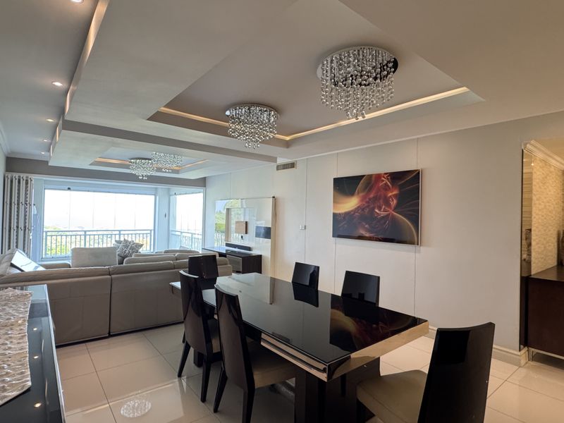 Spacious open-plan living and dining area featuring modern recessed ceilings with crystal chandeliers, tiled flooring, and large sliding doors leading to a balcony.