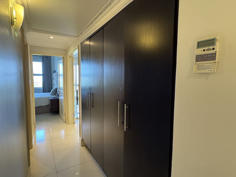 A bright hallway featuring polished tile flooring, extensive built-in storage cupboards, and a wall-mounted climate control panel, leading towards a bedroom.