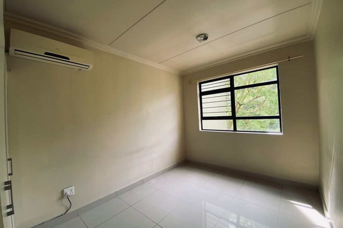 A clean, empty bedroom featuring tiled flooring, a wall-mounted air conditioning unit, and a window with security bars.
