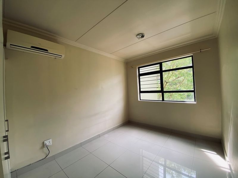 A clean, empty bedroom featuring tiled flooring, a wall-mounted air conditioning unit, and a window with security bars.