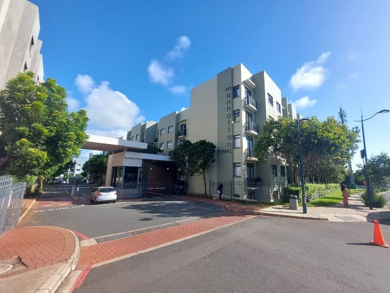 Exterior view of the Manhattan Mews apartment complex, featuring a secure gated entrance, paved driveway, and multi-story residential building with balconies.