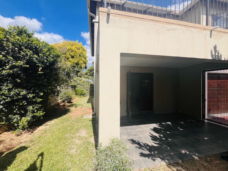 A covered patio area with tiled flooring, adjacent to a private garden space, featuring a security gate and upper-level balcony railing.