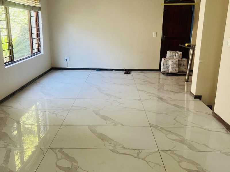A bright, vacant living area featuring modern large-format marble-look floor tiles, recessed lighting, and large windows, currently undergoing minor finishing touches.
