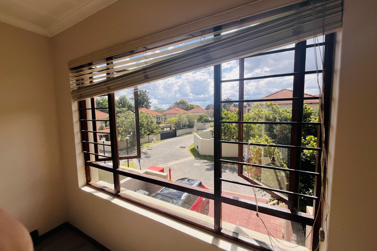 A bright bedroom featuring a large window with security bars and venetian blinds, offering a view of the surrounding residential estate.