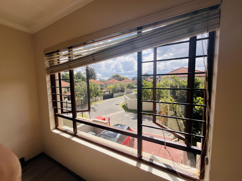 A bright bedroom featuring a large window with security bars and venetian blinds, offering a view of the surrounding residential estate.